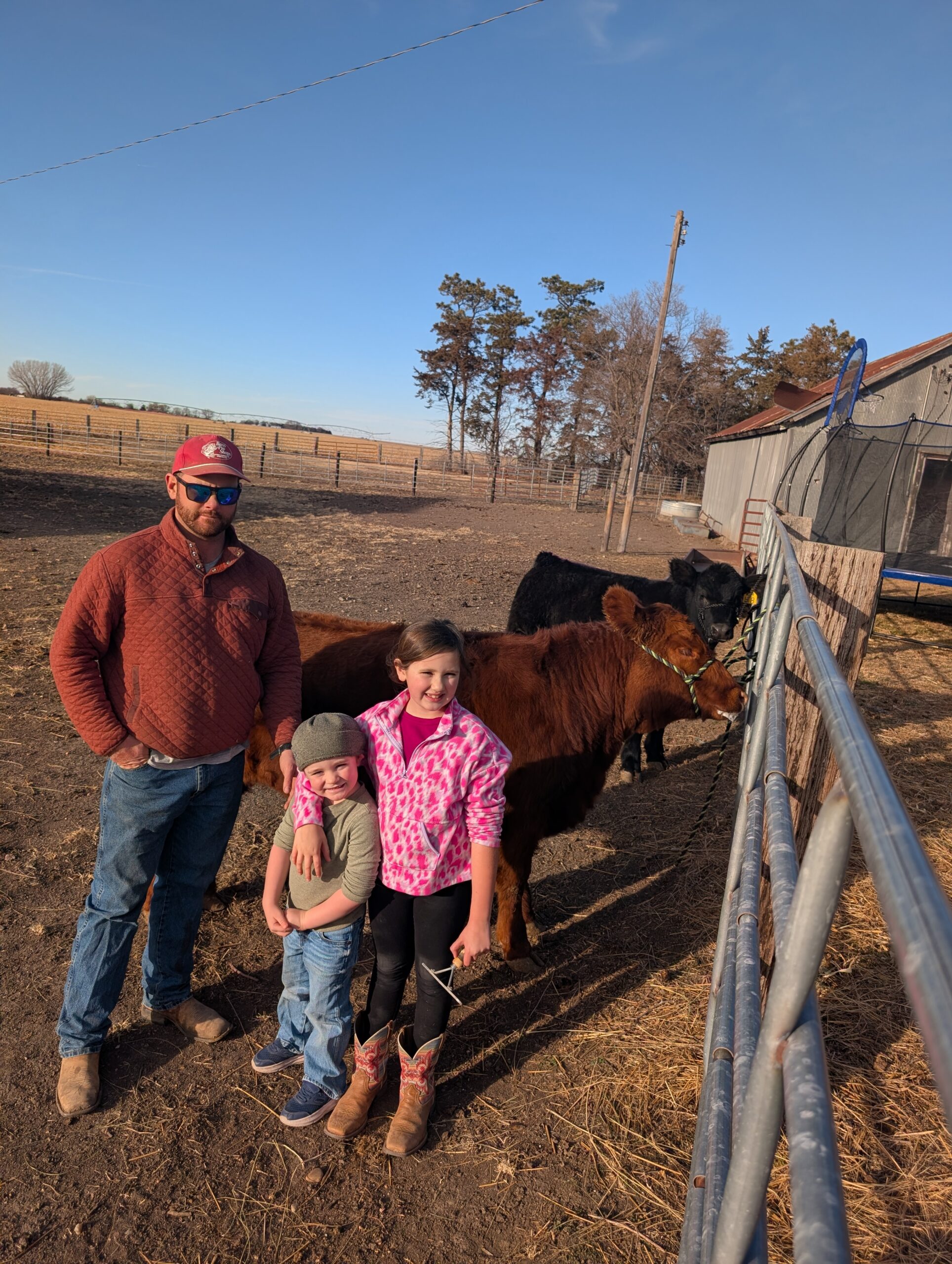 A farmer and two children standing in a pasture with cows, showcasing family farming life. The scene includes a clear blue sky and a barn in the background, emphasizing rural agriculture and livestock care.